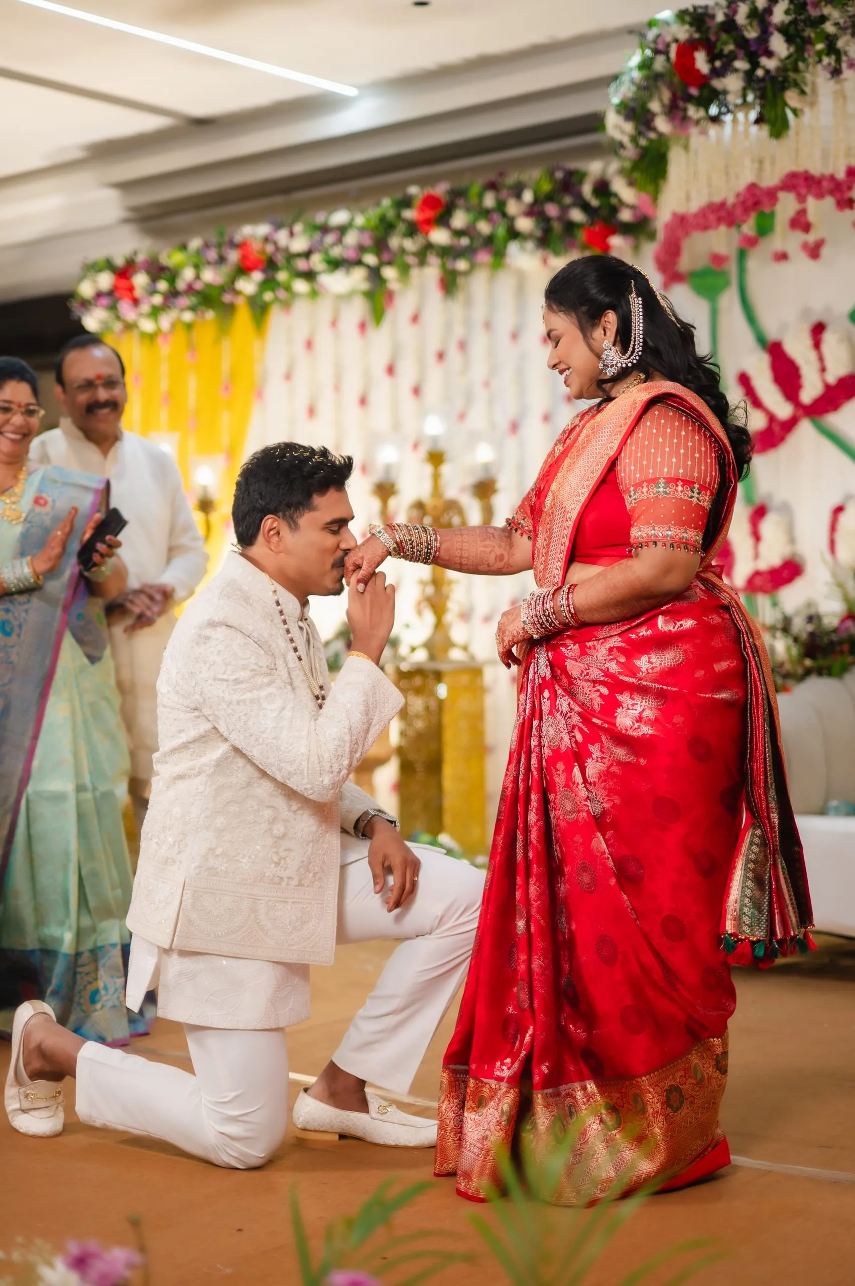 Gautham kneeling to place a ring on Swathi, red saree, flowers behind